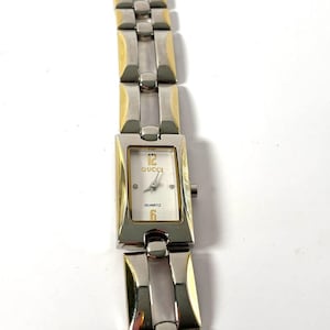 Puede incluir: Un reloj Gucci de cuarzo rectangular con una correa de metal plateado y dorado. La esfera del reloj es blanca con números negros y el logotipo de Gucci. El reloj tiene un bisel plateado y detalles dorados.