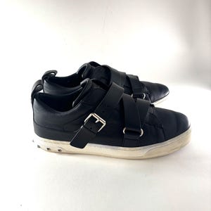 May include: Black leather sneakers with two wide straps and silver buckles. The shoes have a white sole with small, silver studs around the base. The sneakers are set against a white background.