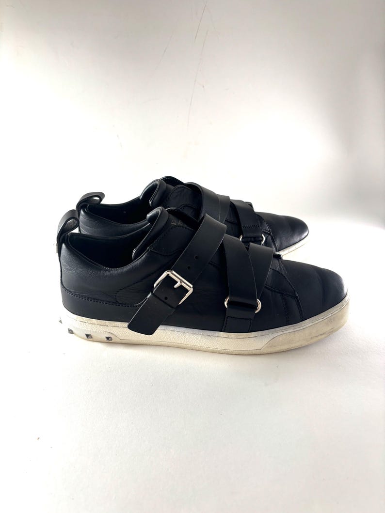 May include: Black leather sneakers with two wide straps and silver buckles. The shoes have a white sole with small, silver studs around the base. The sneakers are set against a white background.