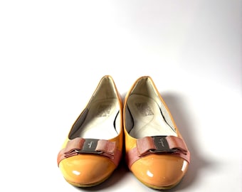 Peut inclure: Une paire de ballerines en cuir verni orange avec un détail de nœud. Les chaussures ont un bout arrondi et un petit talon. Le nœud est de couleur rose clair avec un accent métallique argenté.