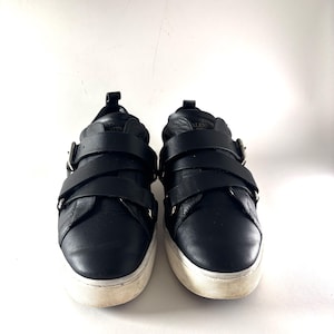 May include: Pair of black leather sneakers with two wide straps across the top, secured by silver buckles. The sneakers have a white sole with a worn appearance. The shoes are set against a plain white background.