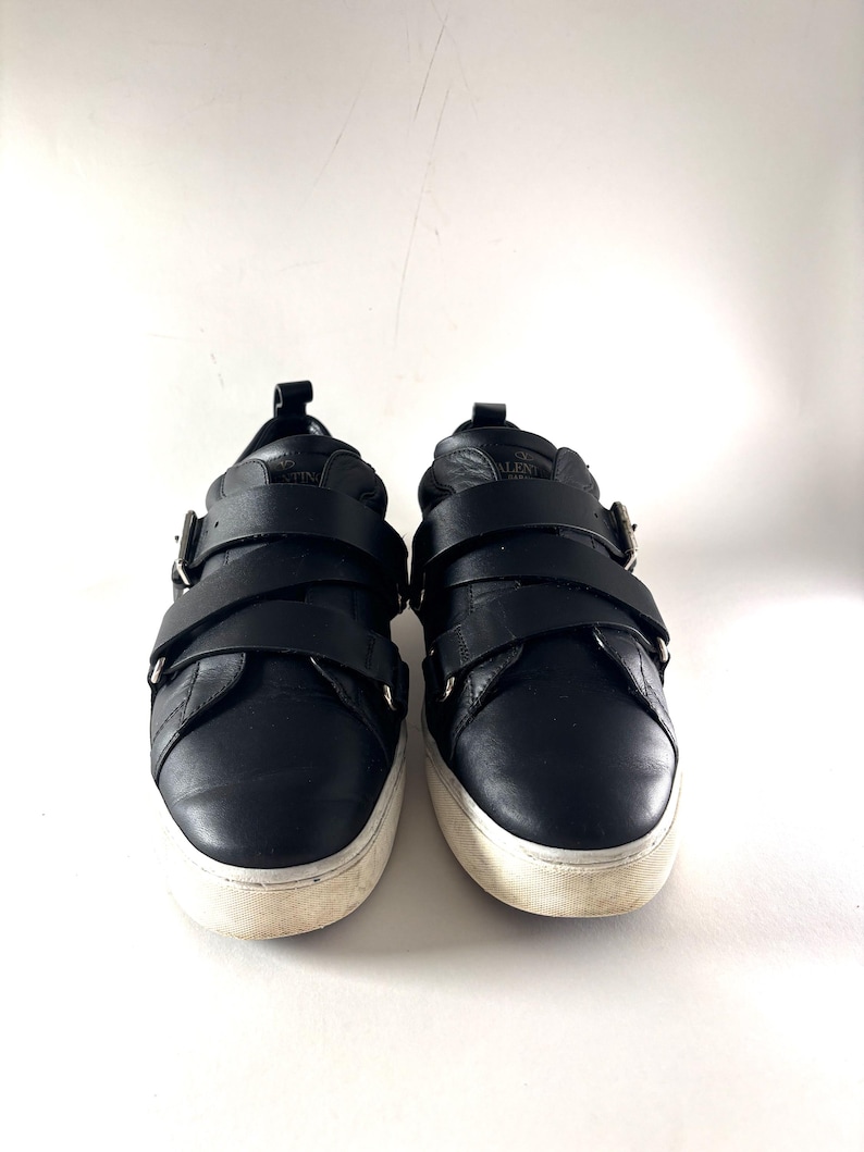 May include: Pair of black leather sneakers with two wide straps across the top, secured by silver buckles. The sneakers have a white sole with a worn appearance. The shoes are set against a plain white background.