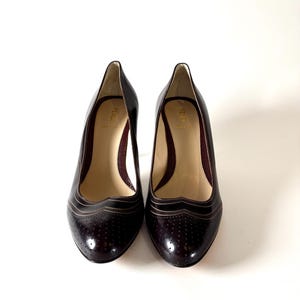 May include: A pair of dark burgundy leather pumps with a pointed toe and a medium heel. The shoes have a perforated design on the toe and a decorative trim. The interior is a light beige color with a burgundy mesh lining. The brand name "FENDI" is visible inside the shoe.