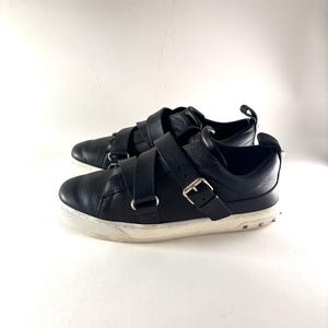 May include: A pair of black leather sneakers with multiple straps and silver buckles. The sneakers have a white sole with small silver studs around the edge. The shoes are set against a white background.