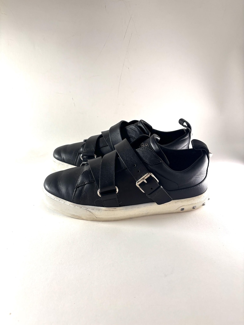 May include: A pair of black leather sneakers with multiple straps and silver buckles. The sneakers have a white sole with small silver studs around the edge. The shoes are set against a white background.
