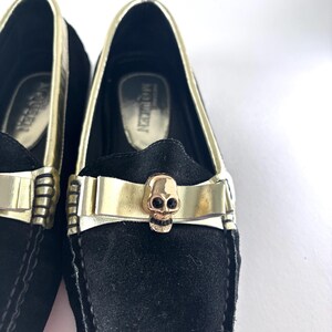 A close-up of the front of a black suede loafer, featuring a gold band with a gold skull emblem on the vamp.