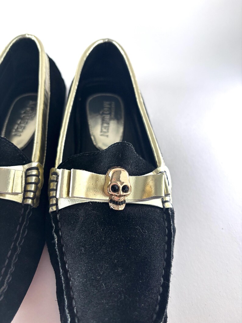 A close-up of the front of a black suede loafer, featuring a gold band with a gold skull emblem on the vamp.