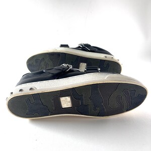 May include: Black leather sneakers with silver-tone studs around the white sole. The shoes have a buckle closure and a textured rubber sole. The soles have a unique pattern. The shoes are displayed on a white background.