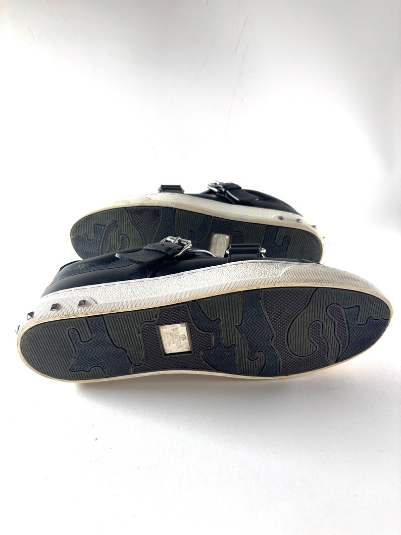 May include: Black leather sneakers with silver-tone studs around the white sole. The shoes have a buckle closure and a textured rubber sole. The soles have a unique pattern. The shoes are displayed on a white background.
