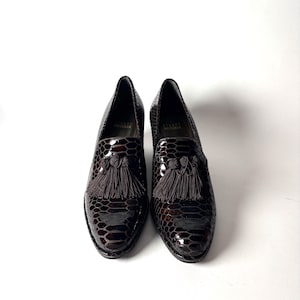 May include: A pair of dark brown leather loafers with a snakeskin pattern. Each shoe has a tassel detail and a glossy finish. The shoes are set against a white background.