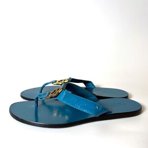 May include: A pair of blue leather flip-flops with gold-colored hardware. The sandals have a glossy finish and a black sole. The Gucci logo is visible on the straps.