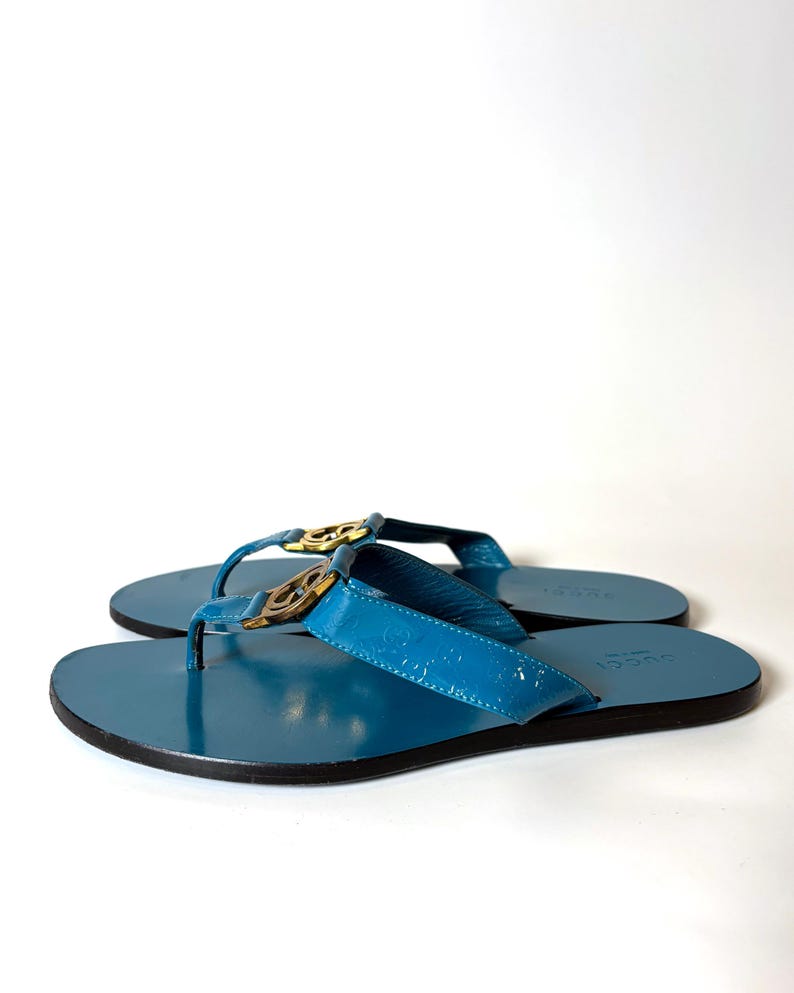 May include: A pair of blue leather flip-flops with gold-colored hardware. The sandals have a glossy finish and a black sole. The Gucci logo is visible on the straps.