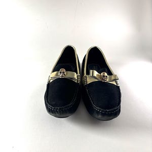 A pair of black suede loafers with gold trim and a gold skull emblem on the vamp, viewed from the front.