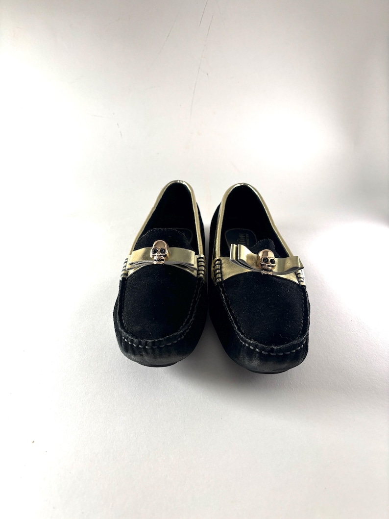 A pair of black suede loafers with gold trim and a gold skull emblem on the vamp, viewed from the front.