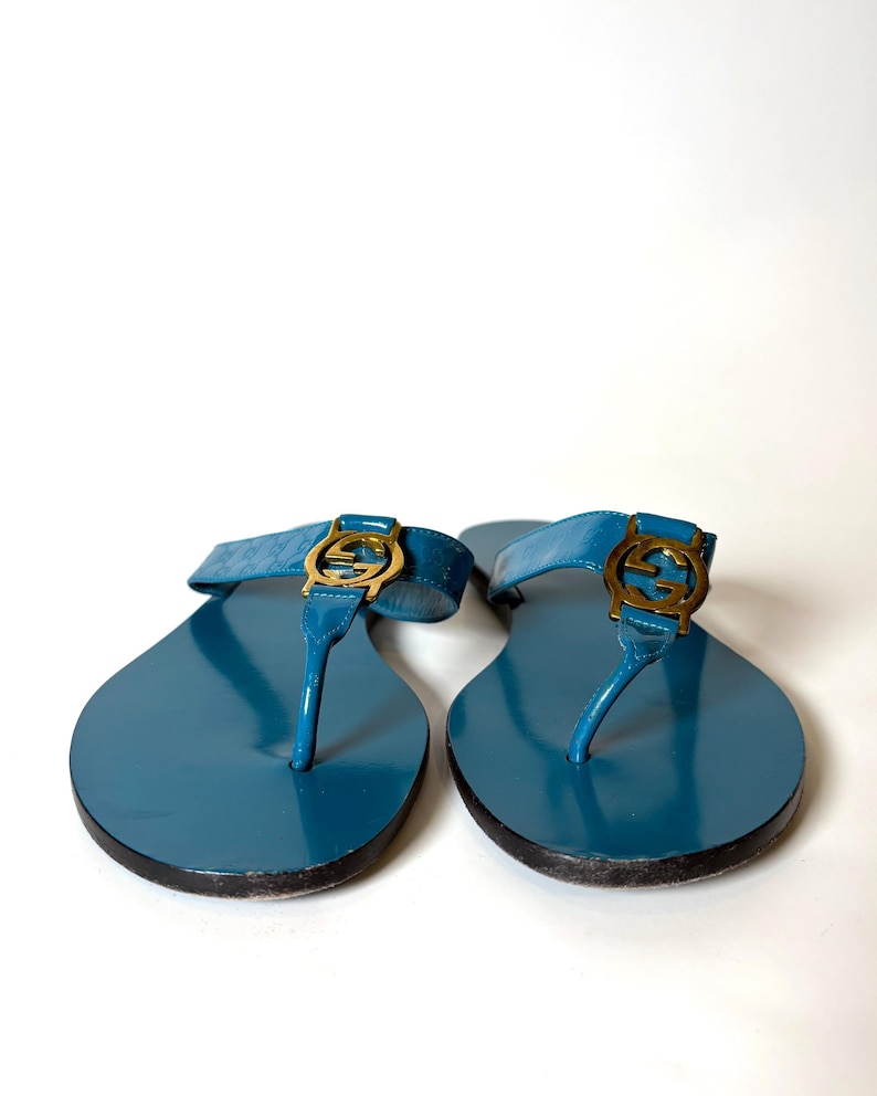 Front view of blue patent leather flat sandals featuring gold-tone interlocking G hardware on a white background.