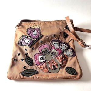 Peut inclure: Un sac à main beige avec un motif décoratif de papillon et de fleur. Le papillon est violet et la fleur est rose et marron. Le sac a une bandoulière en cuir marron et un fermoir en métal argenté.