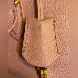 A close-up of a pink leather tag, attached to a gold-toned ring, with visible stitching and textured leather.