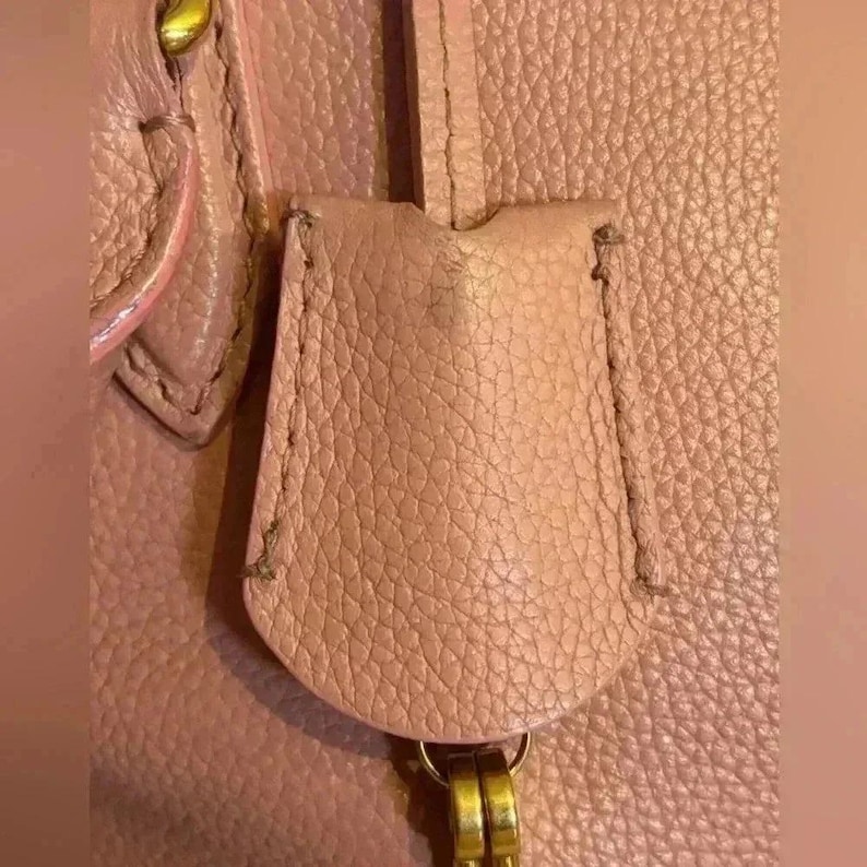 A close-up of a pink leather tag, attached to a gold-toned ring, with visible stitching and textured leather.