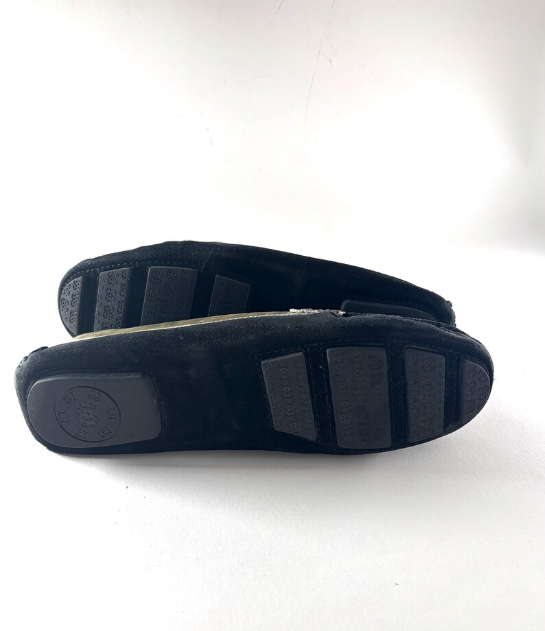 The black rubber soles of two black suede loafers, showing textured grips and a circular logo on one heel.
