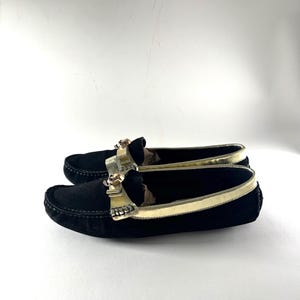 A pair of black suede loafers with gold trim and skull emblems, positioned side-by-side and viewed from a slight angle.