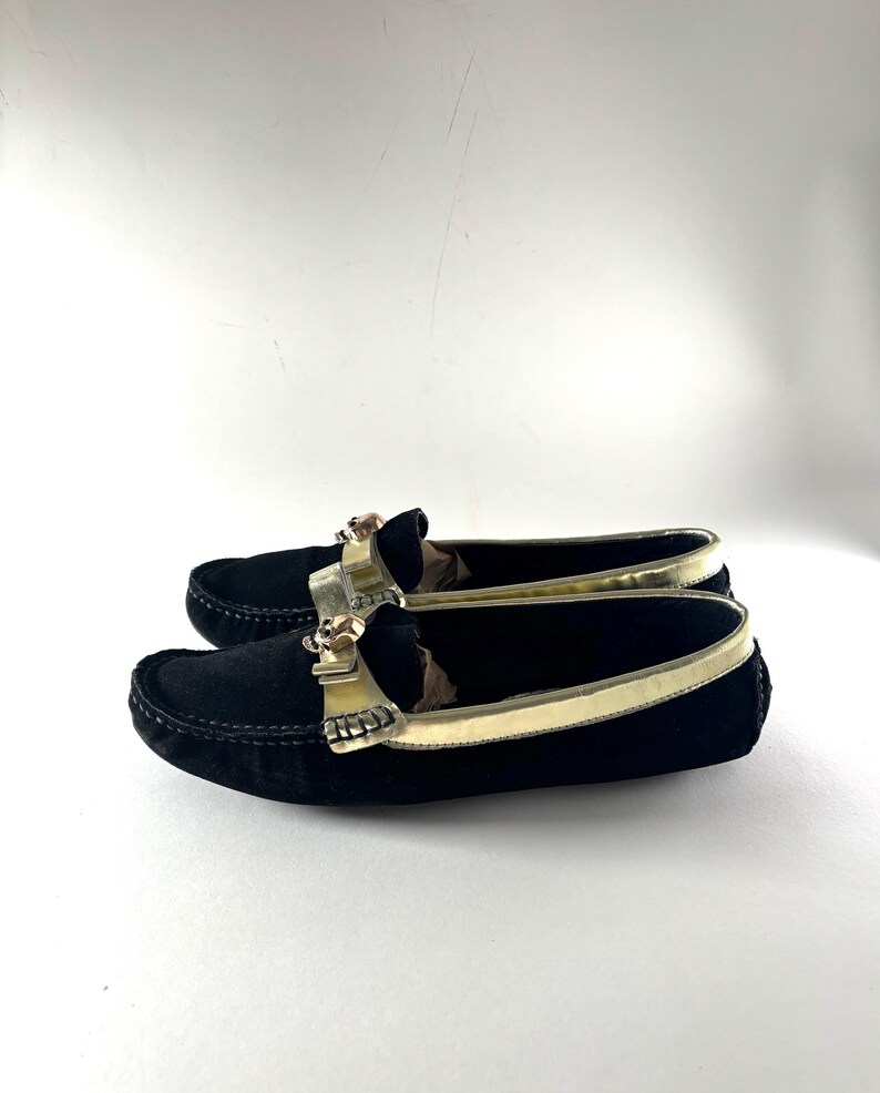 A pair of black suede loafers with gold trim and skull emblems, positioned side-by-side and viewed from a slight angle.