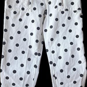 May include: White pants featuring a black polka dot pattern throughout. The pants include two vertical zippered pockets on the front, a button closure, and a small black label on the right pocket area with the white text MJ and two stars.
