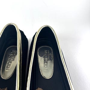 A close-up of the interior of two black suede loafers, showing gold insoles with "Alexander McQueen" branding.