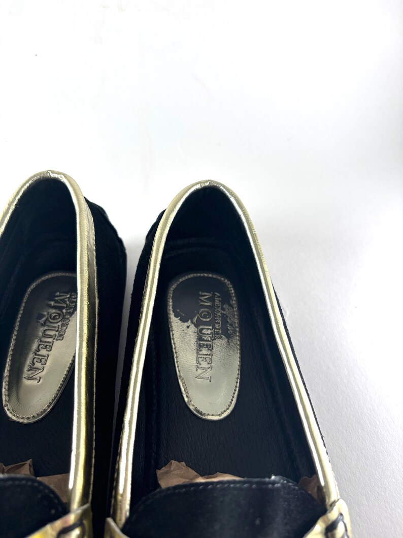 A close-up of the interior of two black suede loafers, showing gold insoles with "Alexander McQueen" branding.
