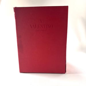 May include: A rectangular red box with the embossed text "VALENTINO GARAVANI" in the center. The box has a slightly distressed appearance, with minor wear and tear visible on the edges.