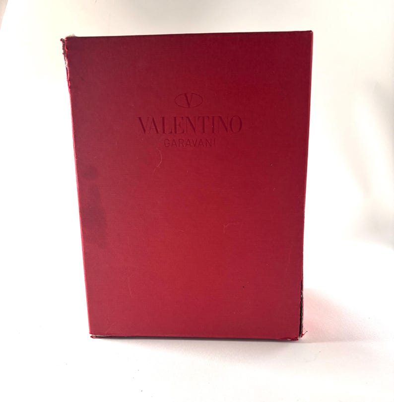 May include: A rectangular red box with the embossed text "VALENTINO GARAVANI" in the center. The box has a slightly distressed appearance, with minor wear and tear visible on the edges.