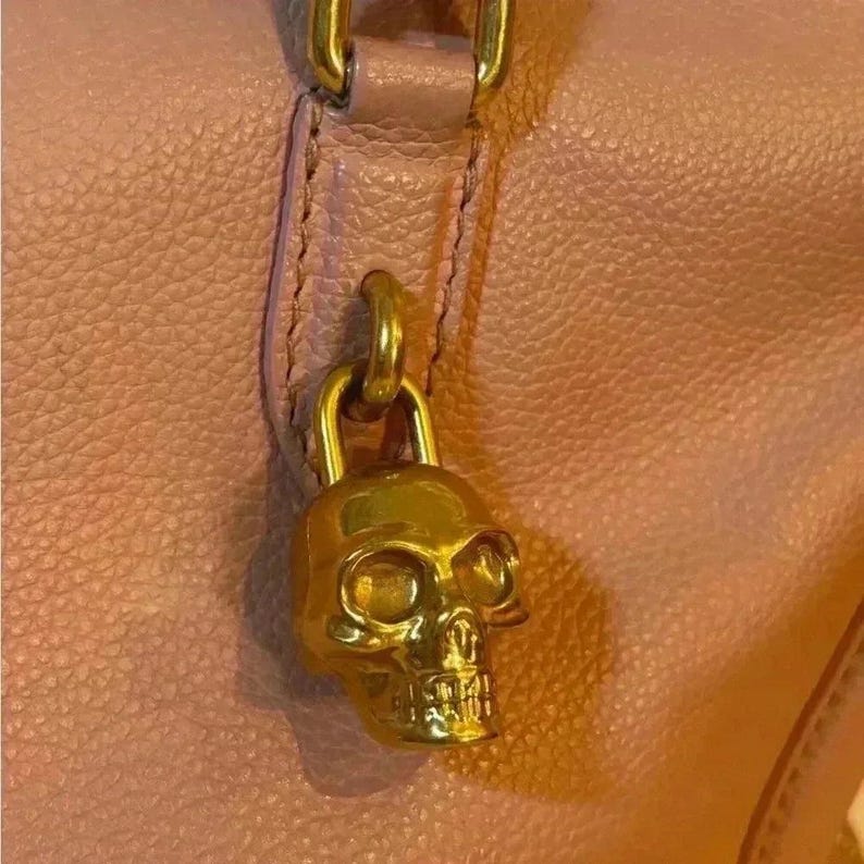 A close-up of a shiny gold skull charm with a ring at the top, attached to a pink leather bag.