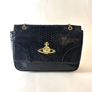 May include: Black handbag with a snakeskin texture and gold-tone chain straps. The bag features a gold-colored emblem on the front. The bag has a rectangular shape and a flap closure.