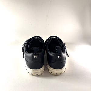May include: A pair of black leather sneakers with silver-tone stud accents on the heel and sole. The shoes have a low-top design with a strap closure. The soles are off-white.