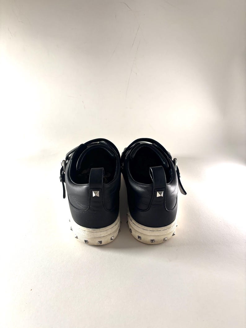 May include: A pair of black leather sneakers with silver-tone stud accents on the heel and sole. The shoes have a low-top design with a strap closure. The soles are off-white.