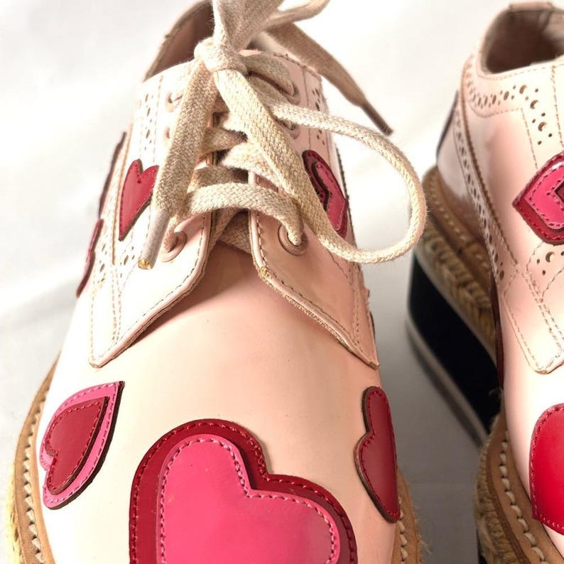 Close-up of the front lacing and layered red and pink heart appliques on the upper of a pink leather oxford shoe.
