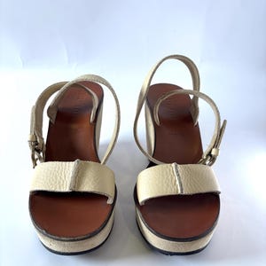 May include: A pair of cream-colored platform sandals with brown soles and ankle straps. The sandals have a chunky heel and a buckle closure. The platform measures approximately 1.5 inches.