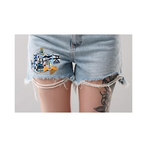 Può includere: Pantaloncini in denim azzurro con orlo sfrangiato e una dettagliata toppa ricamata sulla gamba destra. Il ricamo raffigura una struttura architettonica multicolore con forme e linee geometriche blu, bianche, nere e gialle.