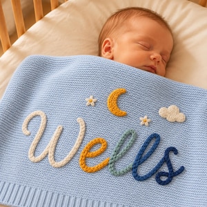 May include: A light blue knitted baby blanket featuring the name Wells in raised, multi-coloured yarn lettering. The design includes a yellow crescent moon, two small white flowers, and a white cloud stitched onto the textured fabric surface.
