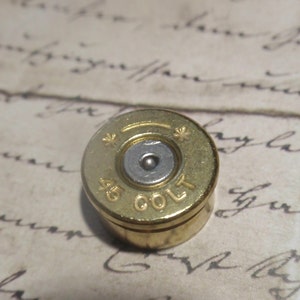 May include: A small, gold-colored metal container with the text "45 COLT" engraved on its side. The container has a circular shape with a silver-colored center and star-shaped designs.