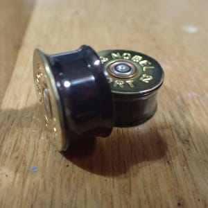 May include: Two cylindrical ear plugs with a flared design, featuring a dark brown body and a metallic gold-colored base. The base is stamped with the text NOBEL SPORT, and a central silver-toned primer is visible on the flat circular end.