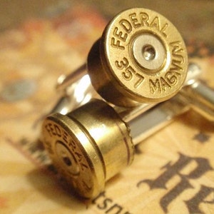 May include: A pair of brass cufflinks made from .357 Magnum bullet casings. The cufflinks have a silver backing and are engraved with "FEDERAL" and "357 MAGNUM". The background is a blurred, patterned surface.