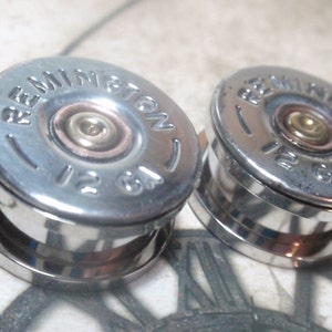 May include: A pair of silver-colored ear gauges with a round, flat top. The top features a silver-colored rim with the words "Remington 12 GA" printed on it. The center of the top is a copper-colored circle with a gold-colored center.