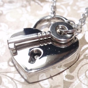 May include: A silver heart-shaped padlock necklace with a key attached. The padlock has a keyhole and a small loop for attaching the chain.