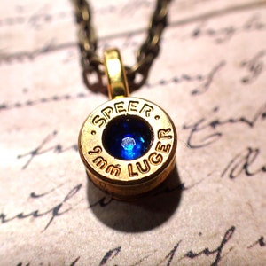 May include: A gold-colored bullet casing necklace with a blue gemstone set in the center. The casing is engraved with "SPEE-R 9mm LUGER".