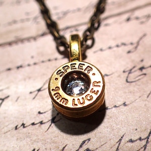 May include: A gold necklace with a bullet casing pendant featuring a clear rhinestone and the inscription "SPEEER 9mm LUGER".