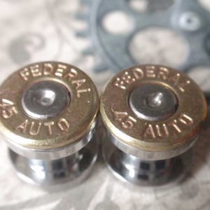 May include: A pair of cufflinks made from 45 Auto bullet casings. The casings are gold-colored with the words "FEDERAL 45 AUTO" inscribed on them. The casings are attached to silver-colored metal bases.