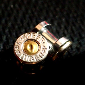 May include: A close-up of a silver bullet casing with the words "SPEED-LUGER" engraved on the side. The bullet casing is on a black fabric background.