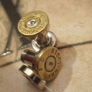 May include: A pair of silver ear gauges with gold-colored bullet casings. The casings have the text "WM9  LUGER  F.C." engraved on them.