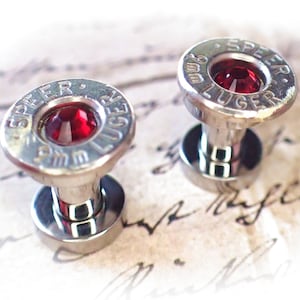 Puede incluir: Un par de gemelos de bala de plata con piedras preciosas rojas. Los gemelos están grabados con "9mm Luger" y "Speer".