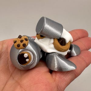 May include: A 3D-printed toy in the shape of a cartoon character, featuring a silver body, white fur, and brown cookie accents. The character has large, expressive eyes and a whimsical design. The toy is held in a hand.